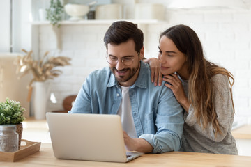 Happy young man and woman hugging, using laptop together, looking at screen, loving couple shopping or chatting online, using internet banking services, reading news in social network