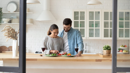 Wall Mural - Happy young loving couple preparing dinner in kitchen at home together, celebrating anniversary, enjoying romantic date, drinking red wine, smiling beautiful wife cutting vegetables for salad