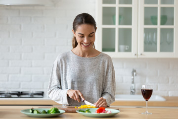 Smiling young woman cutting vegetables for salad on wooden board, using knife, standing in modern kitchen with red wine glass, happy female preparing food for dinner or party alone