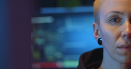 Close up of half face of Caucasian middle-aged woman with short blonde hair looking straight to camera in dark IT office with many screens and data. At night. Female anonymous hacker concept.
