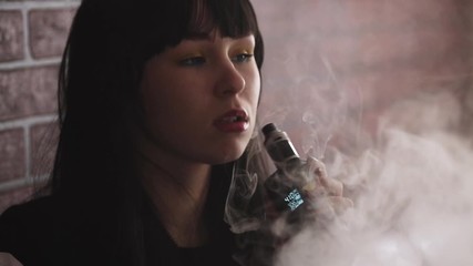 Wall Mural - Vaping teenager. Young pretty white caucasian teenage brunette girl with blue eyes and problem skin smoking an electronic cigarette indoors. Deadly bad habit. Vape activity.