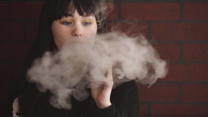 Wall Mural - Vaping teenager. Young pretty white caucasian teenage brunette girl with blue eyes and problem skin smoking an electronic cigarette indoors. Deadly bad habit. Vape activity.