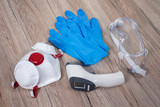 Maska ochronna, rękawiczki jednorazowe, termometr elektroniczny, gogle ochronne. Przedmioty do ochrony przed zarażeniem wirusem leżą na blacie.