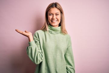 Poster - Young beautiful blonde woman wearing winter wool sweater over pink isolated background smiling cheerful presenting and pointing with palm of hand looking at the camera.