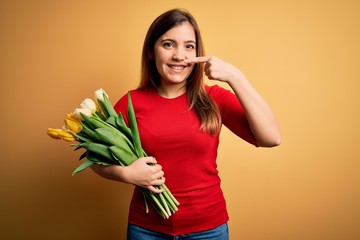 Poster - Young blonde woman holding romantic bouquet of tulips flowers over yellow background Pointing with hand finger to face and nose, smiling cheerful. Beauty concept