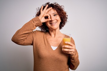 Poster - Middle age curly hair woman drinking healthy glass of orange juice over white background with happy face smiling doing ok sign with hand on eye looking through fingers