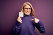 Middle age beautiful blonde woman wearing glasses holding reminder paper with yes message with surprise face pointing finger to himself
