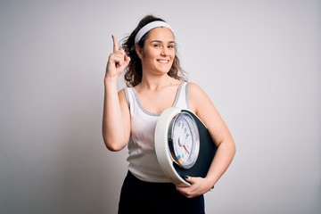 Sticker - Young beautiful woman with curly hair holding weighing machine over white background pointing finger up with successful idea. Exited and happy. Number one.