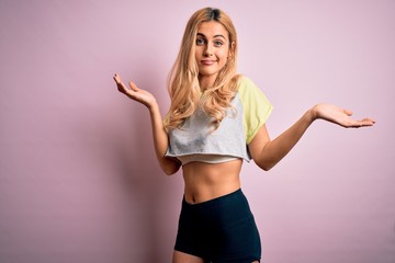 Sticker - Young beautiful blonde sporty woman doing sport wearing sportswear over pink background clueless and confused expression with arms and hands raised. Doubt concept.