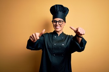 Poster - Young beautiful brunette chef woman wearing cooker uniform and hat over yellow background approving doing positive gesture with hand, thumbs up smiling and happy for success. Winner gesture.