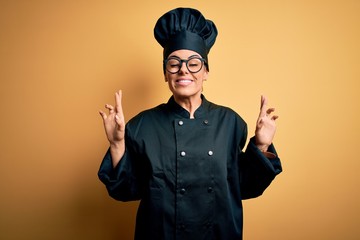 Wall Mural - Young beautiful brunette chef woman wearing cooker uniform and hat over yellow background gesturing finger crossed smiling with hope and eyes closed. Luck and superstitious concept.