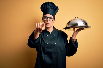 Canvas Print - Young beautiful brunette chefwoman wearing cooker uniform and hat holding tray with dome pointing with finger to the camera and to you, hand sign, positive and confident gesture from the front