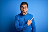 Fototapeta  - Young handsome man with beard wearing casual sweater and glasses over blue background Surprised pointing with finger to the side, open mouth amazed expression.