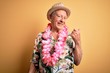 Grey haired senior man wearing summer hat and hawaiian lei over yellow background smiling with happy face looking and pointing to the side with thumb up.