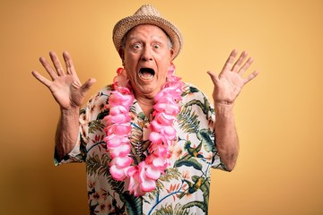 Grey haired senior man wearing summer hat and hawaiian lei over yellow background celebrating crazy and amazed for success with arms raised and open eyes screaming excited. Winner concept