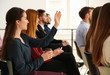 © New Africa - Young woman raising hand to ask question at business training indoors