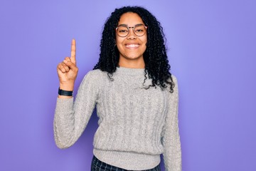 Young african american woman wearing casual sweater and glasses over purple background showing and pointing up with finger number one while smiling confident and happy.