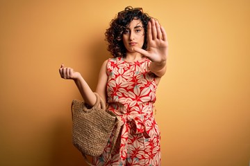 Sticker - Young curly arab woman on vacation wearing floral dress and sunglasses holding wicker bag with open hand doing stop sign with serious and confident expression, defense gesture