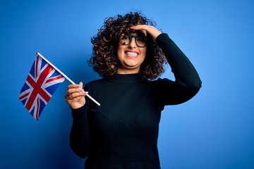 Sticker - Young beautiful curly arab woman wearing glasses holding uk flag over blue background stressed with hand on head, shocked with shame and surprise face, angry and frustrated. Fear and upset