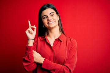 Young beautiful brunette woman wearing casual shirt standing over red background with a big smile on face, pointing with hand and finger to the side looking at the camera.