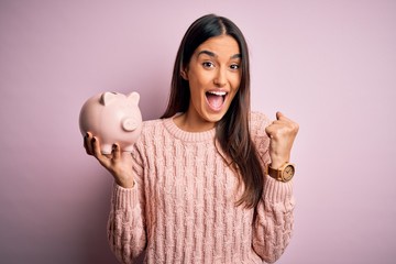 Young beautiful brunette woman holding piggy bank saving money for retirement screaming proud and celebrating victory and success very excited, cheering emotion