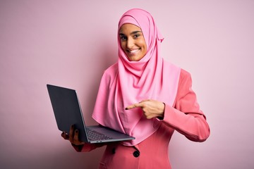 Young beautiful businesswoman wearing pink muslim hijab and business jacket using laptop very happy pointing with hand and finger