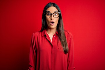 Young beautiful brunette woman wearing casual shirt and glasses over red background afraid and shocked with surprise expression, fear and excited face.