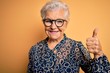 © Krakenimages.com - Senior beautiful grey-haired woman wearing casual shirt and glasses over yellow background doing happy thumbs up gesture with hand. Approving expression looking at the camera showing success.