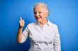 © Krakenimages.com - Senior beautiful woman wearing elegant shirt standing over isolated blue background doing happy thumbs up gesture with hand. Approving expression looking at the camera showing success.