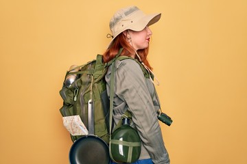 Poster - Young redhead backpacker woman hiking wearing backpack and hat over yellow background looking to side, relax profile pose with natural face and confident smile.