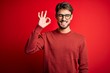 © Krakenimages.com - Young handsome man with beard wearing glasses and sweater standing over red background smiling positive doing ok sign with hand and fingers. Successful expression.