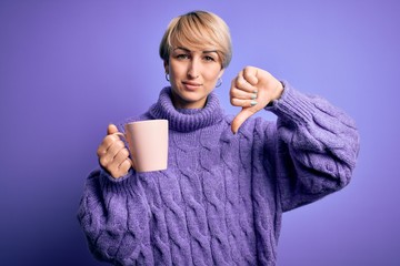 Sticker - Young blonde woman with short hair wearing winter sweater drinking a cup of hot coffee with angry face, negative sign showing dislike with thumbs down, rejection concept