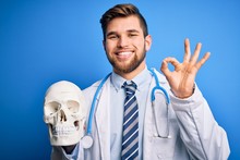 Young Blond Doctor Man With Beard And Blue Eyes Wearing Coat And Stethoscope Holding Skull Doing Ok Sign With Fingers, Excellent Symbol