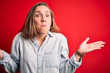 Poster - Young beautiful blonde woman wearing casual denim shirt over isolated red background clueless and confused expression with arms and hands raised. Doubt concept.