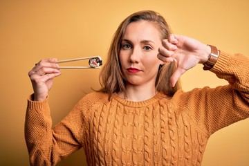 Poster - Young beautiful blonde woman eating sushi using chopsticks over isolated yellow background with angry face, negative sign showing dislike with thumbs down, rejection concept