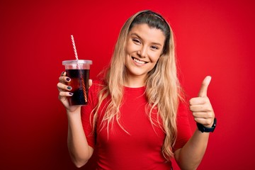 Poster - Young beautiful blonde woman drinking cola fizzy beverage using straw over red background happy with big smile doing ok sign, thumb up with fingers, excellent sign