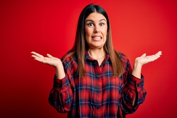 Poster - Young beautiful woman wearing casual shirt over red background clueless and confused expression with arms and hands raised. Doubt concept.