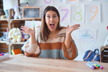 Canvas Print - Young beautiful teacher woman wearing sweater and glasses sitting on desk at kindergarten celebrating crazy and amazed for success with arms raised and open eyes screaming excited. Winner concept