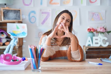 Young beautiful teacher woman wearing sweater and glasses sitting on desk at kindergarten smiling in love showing heart symbol and shape with hands. Romantic concept.