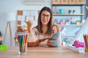 Canvas Print - Young beautiful teacher woman wearing sweater and glasses sitting on desk at kindergarten smiling with happy face looking and pointing to the side with thumb up.