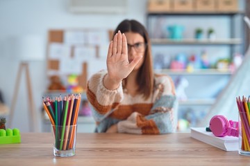 Poster - Young beautiful teacher woman wearing sweater and glasses sitting on desk at kindergarten doing stop sing with palm of the hand. Warning expression with negative and serious gesture on the face.