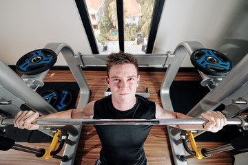 Handsome young strong man looking up when sitting on bench lifting heavy barbell, view from above