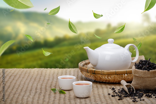 Warm cup of tea with teapot, flying green tea leaves in the air and dried herbs on the bamboo mat at morning in plantations background, Organic product from the nature for healthy with traditional