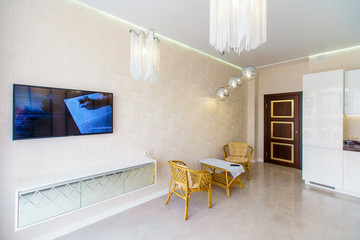 Wall Mural - Kitchen room with a fresh renovation in beige tones. White kitchen set with kitchen appliances. TV, sofa and wicker furniture-table and two chairs