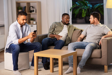 Wall Mural - technology, leisure and people concept - man using smartphone and friends talking at home at night