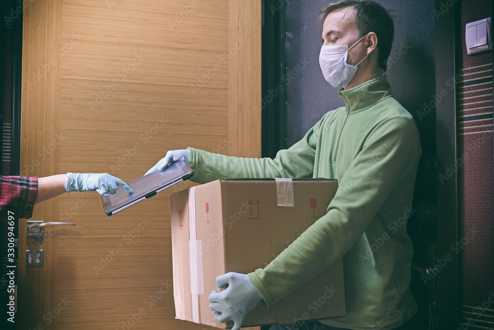 Courier in protective mask delivers parcel to client, client in medical gloves signs on tablet. Delivery service under quarantine, disease outbreak, coronavirus covid-19 pandemic conditions. - obrazy, fototapety, plakaty 