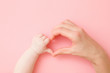 Heart shape created from infant and young mother hands on light pink table background. Pastel color. Lovely emotional, sentimental moment. Love, happiness and safety concept. Closeup. Top down view.