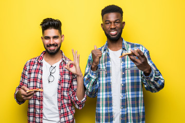 Two mixed race men holding pizza and show thumbs up gesture isolated on yellow background