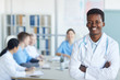 © Seventyfour - Waist up portrait of young African-American doctor smiling at camera while standing with arms crossed against medical conference background, copy space