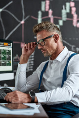 Wall Mural - Smartness is in being sure. Close up of middle-aged caucasian trader looking focused while sitting in front of computer monitor. Blackboard full of charts and data analyses in background. Side view